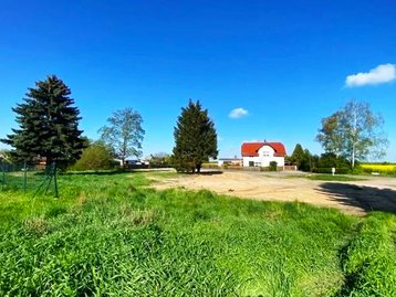 Eine ländliche Szenerie mit einer grünen Wiese im Vordergrund und einem freigeräumten, sandigen Grundstück in der Mitte. Im Hintergrund steht ein weißes Haus mit rotem Dach, umgeben von Bäumen und einer blauen Himmel mit wenigen Wolken.
